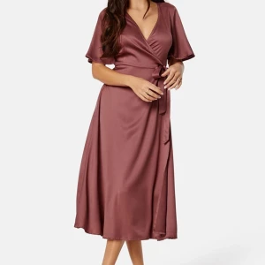 Mörkrosa omlottklänning - Säljer en superfin mörkrosa omlottklänning med knytband i midjan. Klänningen har korta ärmar och en v-ringad design som ger en elegant look. Perfekt för både vardag och fest!