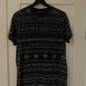 Mönstrad t-shirt från ASOS - Säljer en snygg mönstrad t-shirt från ASOS i storlek M. Den är svart med ett coolt mönster i grått och lite rött. Perfekt för en avslappnad stil. T-shirten är kortärmad och gjord i ett mjukt material.