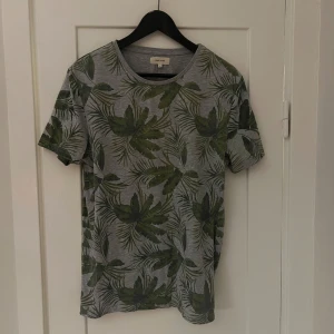 Grå t-shirt med grönt bladmönster från River Island - Säljer en snygg grå t-shirt från River Island med ett coolt grönt bladmönster. Perfekt för en avslappnad stil. T-shirten är kortärmad och i bomull, vilket gör den superbekväm att ha på sig. Passar perfekt till både jeans och shorts!