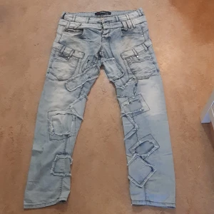 Ljusa jeans med patchwork och slitningar - Säljer ett par unika ljusa jeans från Cipo & Baxx. De har en cool patchwork-design med flera slitningar och detaljerade fickor. Perfekta för en avslappnad och edgy stil. Passar bra till både vardag och fest!