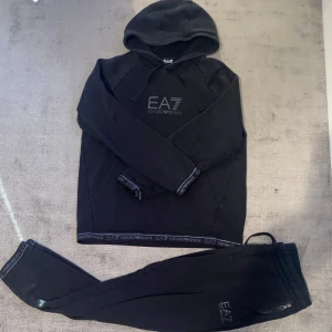 Svart hoodie från Emporio Armani - Säljer en stilren svart Tracksuit från Emporio Armani. Den har en klassisk design med EA7-loggan tryckt på bröstet och ribbade muddar med märkesdetaljer. Perfekt för en casual look eller träning. Superbekväm och snygg! 
