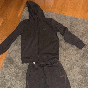 Mörkgrå hoodie, byxor  från BOSS - Snygg mörkgrå hoodie och byxor från BOSS med dragkedja och huva. Perfekt för en avslappnad stil. Matchande byxor finns också tillgängliga. Storlek M/M nästintill oanvända! Kan tänka bytas mot Palm angels tracksuit 