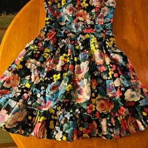 Blommig klänning från D&G - Säljer en färgglad blommig klänning från D&G. Den har en vacker volangdetalj och är perfekt för sommarens festligheter. Klänningen är ärmlös och har en kort längd som ger en lekfull känsla.