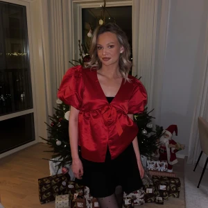 Röd blus med puffärmar - Snygg röd blus med puffiga kortärmar och en stor rosett framtill. Perfekt för festliga tillfällen och ger en elegant touch till din outfit. Blusen har en v-ringad design och är tillverkad i ett glansigt material. Använd en gång, på julafton så i otroligt bra skick 🌟❤️‍🔥 nypris 400kr