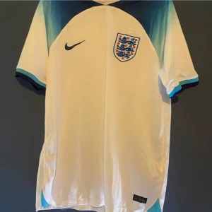 England fotbollströja från Nike - Snygg England fotbollströja från Nike med korta ärmar och ett stilrent märke på bröstet. Tröjan har en cool blå gradientdetalj vid axlarna och är perfekt för fotbollsfans.