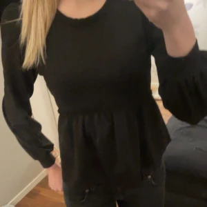 Svart blus med volangdetalj - Snygg svart blus med volangdetalj i midjan och långa ärmar. Perfekt för en stilren look.