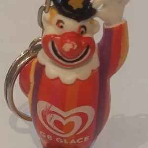 Nyckelring med GB Glace clown - Färgglad nyckelring med en clownfigur från GB Glace. Clownen har en röd och orange dräkt med vita detaljer och en svart hatt. Perfekt för att ge en nostalgisk touch till dina nycklar.