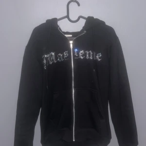 Svart hoodie med glittrig text och motiv - Cool svart hoodie med dragkedja och huva. Framsidan har glittrig text och baksidan ett stort glittrigt motiv med en mask och kort. Perfekt för att sticka ut i mängden!