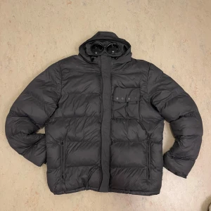 Grå C.P. Company jacka - Vintage C.P. Company puffer. Färgen är gunmetal. Glasögonen går att göma i luvan. Säljer den nytvättad. Storlek XL men passar L. Finns några defekter men ger ett gott helthetsintryck.