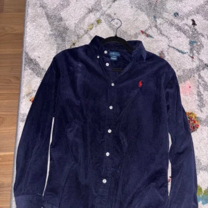 Mörkblå manchester skjorta från Ralph Lauren - Säljer en snygg mörkblå manchester skjorta från Ralph Lauren med klassisk krage och knappar framtill. Den har en broderad röd logga på bröstet och långa ärmar. Perfekt för en stilren look! Storleken är ungdom 16-18 men sitter i runt xs-s
