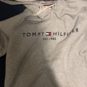 Grå hoodie från Tommy Hilfiger - Säljer en stilren grå hoodie från Tommy Hilfiger med logotyp framtill. Tillverkad i ekologisk bomull och har en klassisk passform med justerbar huva och ribbade muddar.