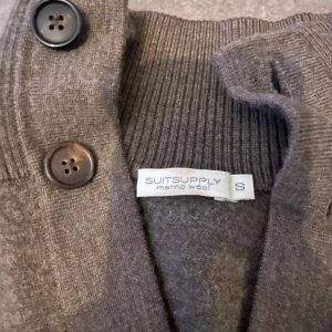 Brun kofta från Suitsupply - Snygg grå kofta från Suitsupply i merinoull. Den har knappar framtill och ribbade detaljer vid ärmslut och nederkant. Perfekt för en stilren look. Den är liten i storleken skulle jag säga, jag är 179-180 och sitter ganska litet och tajt. Annars är den i jätte bra skick inga defekter🙌 nypris runt 2000kr mitt pris 900kr