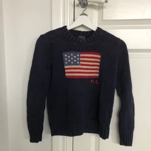 Mörkblå stickad tröja från Polo Ralph Lauren - Säljer en mörkblå stickad tröja från Polo Ralph Lauren med ett stort amerikanskt flaggmotiv på framsidan. Tröjan har långa ärmar och ribbstickade muddar vid ärmslut och nederkant. Perfekt för en avslappnad stil.