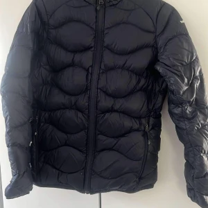 Svart dunjacka från Peak Performance - Snygg svart dunjacka från Peak Performance med quiltad design och dragkedja framtill. Jackan har långa ärmar och en hög krage med R&D-logga på baksidan. Perfekt för kyliga dagar. Finns en liten reva som är limmad bak. Se bild 3
