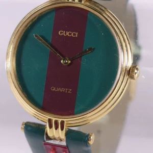 Gucci klocka med guldtonad boett - Elegant Gucci klocka med en guldtonad boett och en unik urtavla i grönt och rött. Klockan har en stilren design med svart och rött armband, perfekt för den modeintresserade.