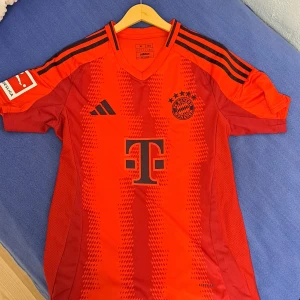 Bayern munchen tröja - Säljer en röd fotbollströja från Adidas med korta ärmar och svarta detaljer. Tröjan har klubbmärke och sponsorlogga på framsidan samt tre svarta ränder längs axlarna. Perfekt för fotbollsfans!