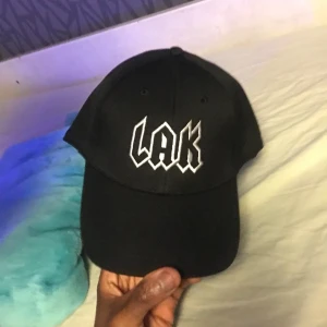 Svart keps med LAK-broderi - Snygg svart keps med vit broderad text 'LAK' framtill. Perfekt för en casual look och att skydda mot solen. Justerbar passform för optimal komfort.