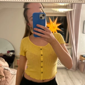 Gul ribbad topp med knappar - Säljer en söt gul ribbad topp med korta ärmar och knappar framtill. Perfekt för en solig dag eller en avslappnad kväll. Toppen är croppad och har en fin passform.