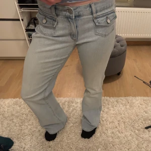 Ljusblå bootcut jeans - Snygga ljusblå bootcut jeans med låg midja och knappar framtill. Jeansen har två framfickor med lock och knappar samt bakfickor med en dekorativ detalj. Perfekta för en avslappnad stil. Näst intill oanvända!