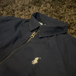 Marinblå jacka från Ralph Lauren - Snygg marinblå jacka från Ralph Lauren med dragkedja och klassisk krage. Jackan har ett broderat logotypmärke på bröstet och är fodrad med ett randigt mönster i blått och vitt. Perfekt för en stilren look. Storlek M 10-12 år! 