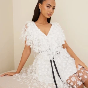 Vit Ida stöstedt klänning med bälte - Snygg vit klänning "Theresa"i storlek 38 från Ida Sjöstedt med blommigt mönster med silvriga trådar och v-ringad design. Den har korta ärmar och ett svart knytbälte i midjan för en snygg silhuett. Perfekt för sommarens festligheter, student eller annan avslutning. Nypris 4600kr. Endast använd en gång och är i nyskick utan defekter.