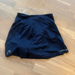 Mörkblå träningskjol från Head - Säljer en mörkblå kjol från Head, perfekt för sportiga aktiviteter mer exakt tennis och padel etc. Den har en stilren design med en liten slits framtill och är tillverkad i ett lätt och bekvämt material. Den är helt ny och nypris ligger på runt 500kr