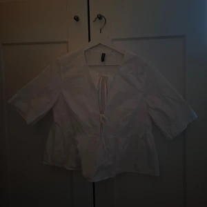 Vit blus från H&M Divided - Säljer en söt vit blus från H&M Divided med knytning framtill. Blusen har trekvartsärmar och en lätt volangdetalj vid midjan. Perfekt för en stilren och avslappnad look.