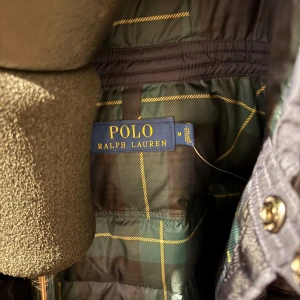 Mörkblå dunjacka från Polo Ralph Lauren - Säljer en snygg mörkblå dunjacka från Polo Ralph Lauren. Jackan har en quiltad design med dragkedja och knappar framtill. Den har en huva och praktiska fickor med lock. Perfekt för kyliga dagar. Köpt för 3700 kvalitet 10/10