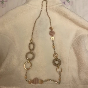 Långt halsband med gulddetaljer - Unikt långt halsband med en mix av guldfärgade och randiga detaljer. Halsbandet har stora cirkulära länkar och pärlor i olika storlekar. Perfekt för att ge en extra touch till din outfit.