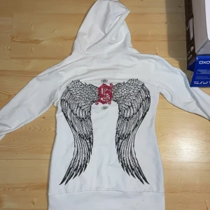 Vit hoodie från Sinful med vingar och rhinestones - Säljer en vit v-ringad hoodie från Sinful med vingar på ryggen med rhinestones. Inga rhinestones fattas.