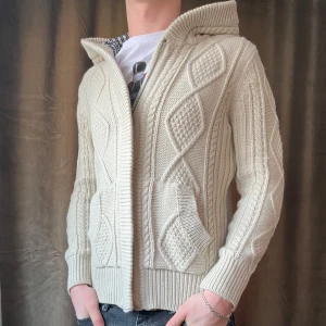 Stickad Cardigan/Hoodie - Säljer en snygg beige stickad hoodie/cardigan. Den har ett klassiskt flätmönster och ribbade muddar. Huvan är fodrad med ett rutigt tyg för extra stil. Perfekt för kyliga dagar! Vid minsta fråga eller fundering hör av er🤝