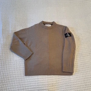 Beige tröja från Calvin Klein - Snygg beige tröja från Calvin Klein med rund hals och ribbade muddar. Den har en stilren design med en svart CK-logga på ärmen. Perfekt för en avslappnad och trendig look.