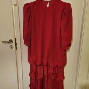 Röd klänning från Claudia - Elegant röd klänning från Claudia med plisserad nederdel och trekvartsärmar. Perfekt för höst och vinter med sin djupa färg och stilrena design. Klänningen har en lös passform och en dekorativ rosett vid midjan.