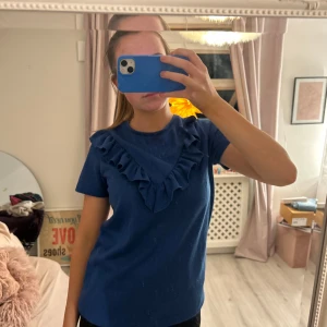Blå t-shirt med volangdetalj - Säljer en blå t-shirt med volangdetalj vid halsringningen. T-shirten har korta ärmar och en normal passform. Perfekt för en stilren och bekväm look.