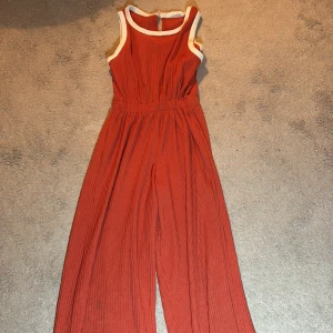 Röd ärmlös jumpsuit från Zara - Säljer en snygg röd ärmlös byxdress från Zara med vita kanter. Byxdressen har en ribbad struktur och vida ben. Perfekt för en stilren look!