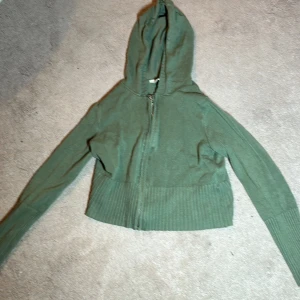 Grön hoodie från H&M - Säljer en grön hoodie från H&M med dragkedja framtill. Tröjan har långa ärmar och en mysig huva, perfekt för kyliga dagar. Ribbstickad nederkant och ärmslut ger en skön passform.
