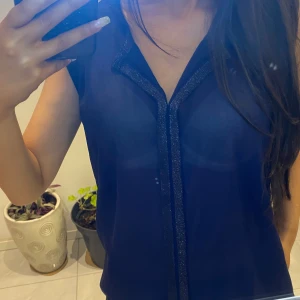 Mörkblå ärmlös blus med glittrig detalj - Snygg mörkblå ärmlös blus med en glittrig kant längs V-ringningen. Perfekt för en stilren look.