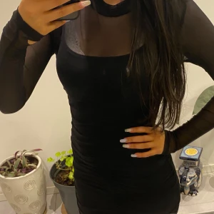 Svart klänning med två delar ❤️ - Snygg svart klänning med transparenta meshdetaljer upptill och långa ärmar. Perfekt för en kväll ute eller en speciell tillställning. Klänningen har en tight passform som framhäver figuren.