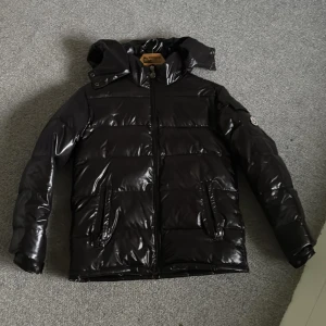 Svart glansig vinter jacka - Snygg svart pufferjacka med glansig finish och dragkedja framtill. Jackan har en praktisk huva och två fickor med knappar. Perfekt nu på vintern!  ALLDRIG ANDVÄND!