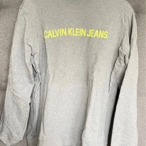 Grå tröja från Calvin Klein Jeans - Säljer en stilren grå tröja från Calvin Klein Jeans med gul text på bröstet. Tröjan har långa ärmar och en rund halsringning. Perfekt för en avslappnad look.