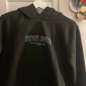 Svart hoodie från Teddy Smith - Cool svart hoodie från Teddy Smith med texten 'Street Culture' och en blå ros på ryggen. Framsidan har en stilren logga i blått och vitt. Perfekt för en avslappnad streetwear-look.