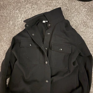 Svart overshirt från Selected Homme - Snygg svart overshirt från Selected Homme med knappar framtill och två bröstfickor. Perfekt för lager-på-lager-stil och passar till många olika outfits. Ända defekten är att knappen på en arm har lossnat men går att fixa enkelt med sy maskin