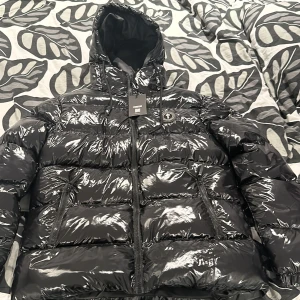 Svart pufferjacka från Trapstar - Snygg svart pufferjacka från Trapstar med glansig finish och huva. Jackan har dragkedja och märkets logga på bröstet. Perfekt för kyliga dagar.