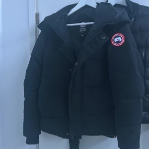 Svart dunjacka från Canada Goose - Säljer en svart dunjacka från Canada Goose med huva och dragkedja. Perfekt för kalla vinterdagar. Jackan har en broderad logotyp på ärmen och är designad för att hålla dig varm och bekväm.använd 1 vinter.