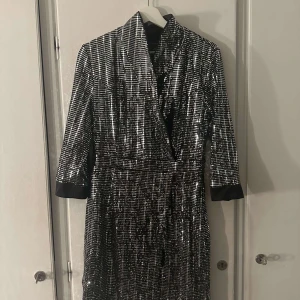 Glittrig klänning med paljetter - Snygg och iögonfallande klänning täckt med silverfärgade paljetter. Den har en omlott-design och trekvartsärmar med svarta detaljer vid ärmsluten. Perfekt för en festlig kväll!