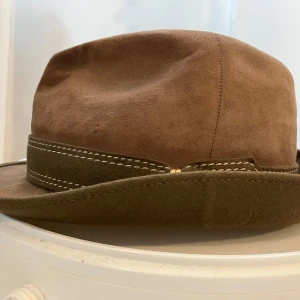 Brun mockahatt med dekorativt band - Snygg brun hatt i mocka med ett dekorativt band runtom. Perfekt för en stilfull vintagelook. Märkt med 'Das Leichen' för hög kvalitet.
