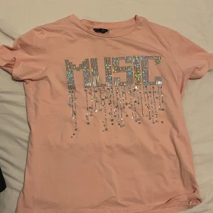 Rosa t-shirt med paljetter från Splash - Snygg rosa t-shirt från Splash med glittrande paljetter som bildar ordet 'MUSIC' på framsidan. Kortärmad och perfekt för en avslappnad stil med en touch av glamour.