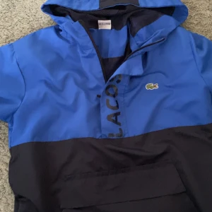 Lacoste windbreaker ( Blå- svart) - Säljer en snygg svart och blå vindjacka från lacoste. Jackan är perfekt för vår och höst! Den har en klassisk Lacoste-logga på bröstet och en större logga på ryggen. 