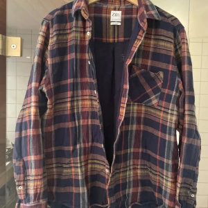 Rutig skjorta från Zara - Säljer en  rutig skjorta från Zara. Den har en mörkblå bas med ränder i orange, gul och beige. Skjortan är i nyskick!