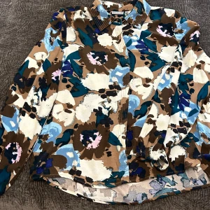 Blommig skjorta med långa ärmar - Säljer en superfin blommig skjorta med långa ärmar. Den har ett färgglatt mönster i brunt, blått, beige och vitt. Perfekt för att liva upp garderoben! Skjortan är i bra skick och passar både till vardags och fest.